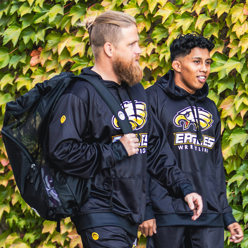 Two men wearing black hoodies with a logo, one carrying a backpack, against a green leafy background.